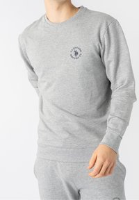 Man som bär en ljusgrå långärmad sweatshirt och matchande sweatpants med en liten U.S. Polo Assn. logotyp på bröstet och byxorna.