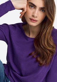 Purple, long-sleeve shirt with a round neckline and soft texture. Worn casually, with a hint of blue denim visible in the frame.