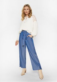 Witte blouse met bloemenkantmouwen, blauwe wijde broek met strik in de taille en beige enkellaarzen met een gestructureerde afwerking.
