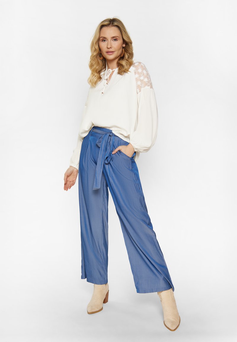 Witte blouse met bloemenkantmouwen, blauwe wijde broek met strik in de taille en beige enkellaarzen met een gestructureerde afwerking.