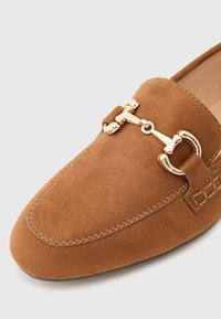Brown suede loafer with gold-tone hardware detail. Features a rounded toe and decorative stitching along the edge. Smooth texture.