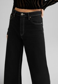 Svarta vida jeans i denim med kontrasterande vit söm, fem fickor och en metallknappstängning i midjan.