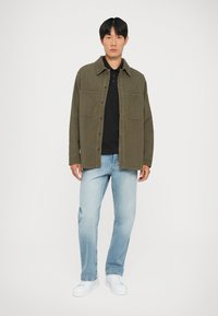 Camisa abotoada verde-oliva com dois bolsos no peito, usada sobre um polo preto. Jeans azul claro e ténis brancos completam o visual.