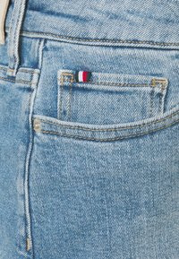 Light blue denim jeans with a textured finish, featuring a small pocket detail with red, white, and navy stripes. Gold stitching visible.