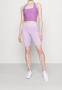 Top corto morado brillante con tirantes gruesos y pantalones cortos de ciclista lilas a juego. Modelo lleva zapatillas blancas; presenta una textura suave y un diseño ajustado.