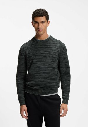 Uomo in piedi di fronte, indossa un maglione a maniche lunghe grigio scuro con trama e pantaloni neri su uno sfondo bianco semplice.