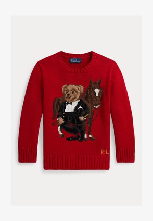 Červený pletený svetr s medvědem ve fraku, který drží otěže a stojí vedle hnědého koně s bílou skvrnou na čele.