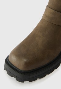 Brown faux leather ankle boot with a rounded toe, textured surface, black rubber sole, and a decorative strap around the upper.