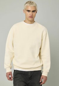 Sweatshirt beige avec un col rond et des poignets côtelés, fabriqué à partir d'un tissu épais et texturé. Porté sur une chemise blanche et associé à un jean noir.
