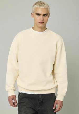 Sweatshirt beige avec un col rond et des poignets côtelés, fabriqué à partir d'un tissu épais et texturé. Porté sur une chemise blanche et associé à un jean noir.
