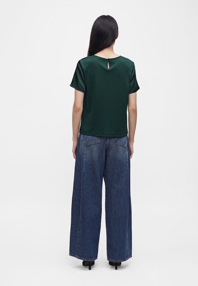Femme aux longs cheveux foncés, de dos, portant un haut vert foncé à manches courtes et un jean bleu à jambes larges avec des talons noirs.
