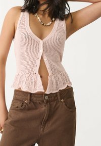 Różowa dzianinowa kamizelka z głębokim dekoltem w kształcie litery V, zapinana na guziki i wykończona falbaną u dołu. W połączeniu z brązowymi spodniami dżinsowymi, podkreślającymi kontrast faktur.