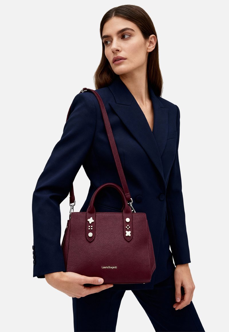 Borsa a mano in ecopelle color bordeaux con doppi manici e tracolla, con borchie decorative e logo dorato sulla parte anteriore.
