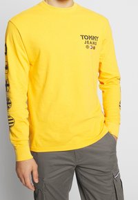 Gelbes Langarm-T-Shirt mit Rundhalsausschnitt, auf dem die Aufschrift "TOMMY JEANS" in Schwarz auf der Brust sowie Logo-Accents an den Ärmeln zu sehen sind.