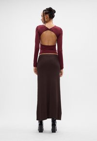 Kobieta z warkoczami nosi burgundową bluzkę z długim rękawem z odkrytymi plecami, brązową maxi spódnicę i czarne botki na obcasie, odwrócona plecami na białym tle.