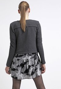 Kvinna med lång hästsvans som bär en mörk texturerad jacka och en svartvitt abstrakt mönstrad kjol, stående vänd bort mot en vit bakgrund.