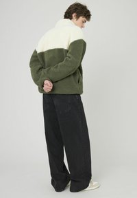 Veste en polaire verte avec une partie supérieure crème, col haut et poches latérales, associée à un jean noir ample et des baskets. Matériau texturé.