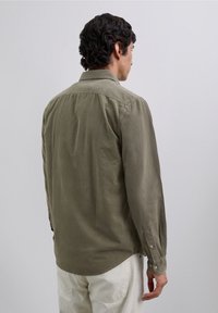 Chemise vert olive à manches longues fabriquée en tissu texturé avec un col boutonné et un ourlet arrondi. Présente des poignets boutonnés et une coupe décontractée.