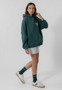 Teal színű, oversized kapucnis pulóver elülső zsebbel, szürke mini szoknya, zöld zoknik és fehér sportcipők. Puha anyag, laza szabás és klasszikus design.