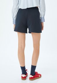Marineblauwe katoenen shorts met een elastische tailleband, van achteren getoond, gecombineerd met rode sneakers en marineblauwe sokken. Simpel en casual ontwerp.