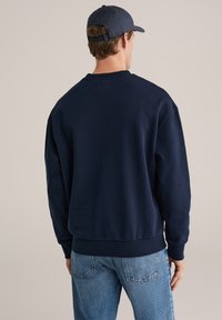 Mornarsko moder pulover iz mehkega blaga z okroglim izrezom in rebrastimi manšetami. Nošen s svetlo modrimi kavbojkami, pogled od zadaj.