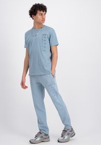 Lichtblauwe katoenen T-shirt en bijpassende sweatpants met zijzakken. Bevat verticale "ALPHA INDUSTRIES" tekst in tonale print. Grijze sneakers.