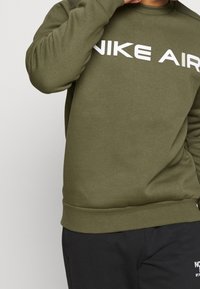 Olivově zelená mikina s kulatým výstřihem a dlouhými rukávy, s výrazným bílým logem "NIKE AIR" vpředu. Měkký materiál, žebrované manžety.