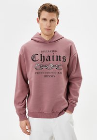 Rosa hoodie med grafisk tryck som visar kedjor och texten "Bryter kedjor Frihet för alla MMXXIV." Mjukt tyg, avspänd passform, ribbade ärmmuddar.
