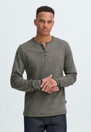 Dunkelgrünes Langarm-Henley-Shirt mit strukturiertem Muster, Rundhalsausschnitt und vier schwarzen Knöpfen an der Knopfleiste, kombiniert mit dunklen Jeans.