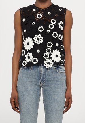 Czarny, bezrękawnik na szydełku z dużymi i małymi białymi kwiatowymi wzorami, noszony z jasnoniebieskimi jeansami z wysokim stanem.