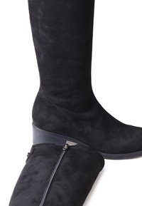 Botas de ante negras hasta la rodilla con un tacón bajo, que cuentan con un zip lateral para facilitar el uso y una superficie suave y texturizada.