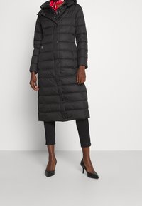 Musta toppatakki hupulla, jossa tikattu kuosi, napit edessä ja pitkät hihat. Päällä mustat legginsit ja korkeakorkoiset kengät.