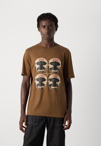 Mann in braunem T-Shirt mit vier Grafiken von Boxhelmen, beschriftet mit "HUGO", und schwarzen Hosen, steht vor einem schlichten, hellen Hintergrund.