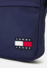 Bolsa de tela azul marino con un parche rectangular que lleva el texto "TOMMY JEANS" y un logotipo en rojo, blanco y azul. Superficie texturizada, costuras resistentes.