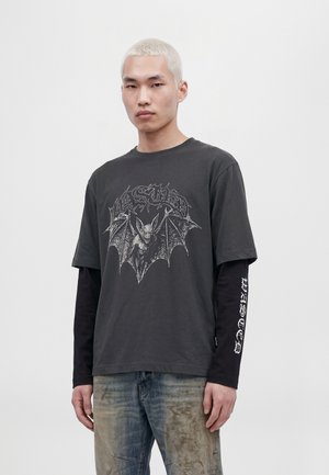 Un modèle aux cheveux courts décolorés porte un haut anthracite à manches longues et superposé, orné d’un motif de chauve-souris, ainsi qu’un jean délavé, sur un fond clair et uni.