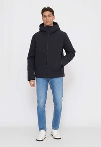 Sort hættejakke med lynlås foran, to sidelommer og en glat tekstur, kombineret med lyseblå jeans og hvide sneakers.