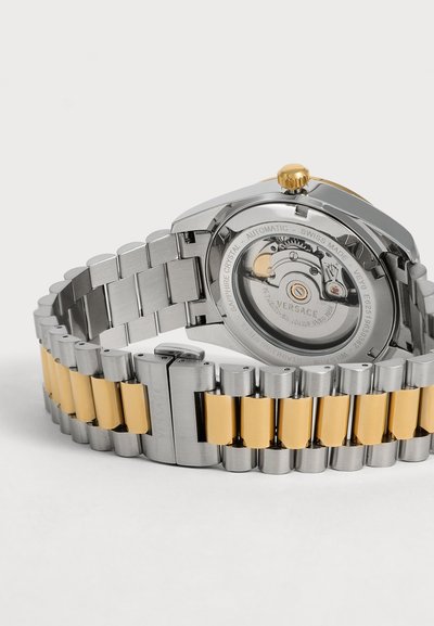 Bracelet de montre en métal bicolore avec des maillons dorés et argentés, montrant le fond avec un mouvement automatique visible et une couronne en or.