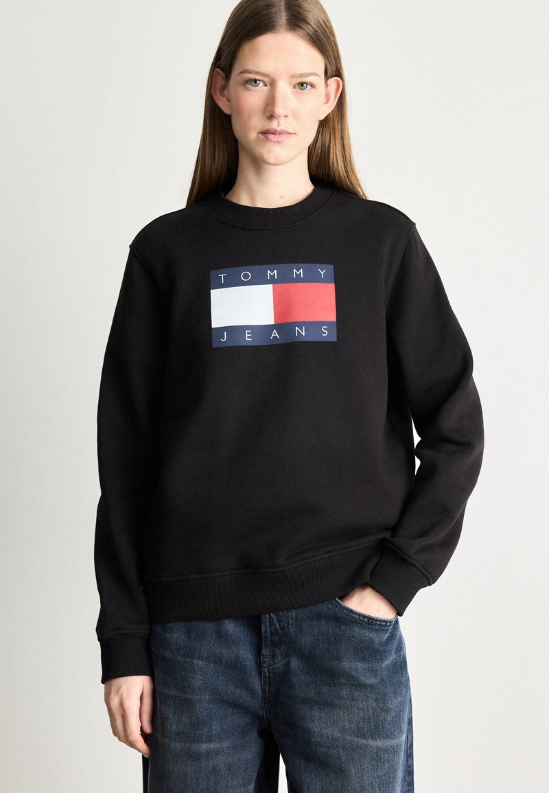 Musta huppari, jossa on ribattu pyöreä kaulus, etupuolella sinisestä, valkoisesta ja punaisesta koostuva suorakaiteen muotoinen logo, jossa lukee "TOMMY JEANS".