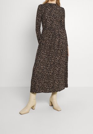 Mujer con un vestido largo de manga larga hasta el tobillo con estampado de leopardo y botas de tacón color crema, de pie contra una pared blanca lisa.