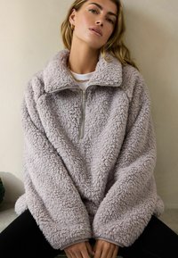 Lys grå fleece pullover jakke med høj krave, kvart lynlås foran og oversized pasform. Har blødt, tekstureret materiale og brede ærmer.