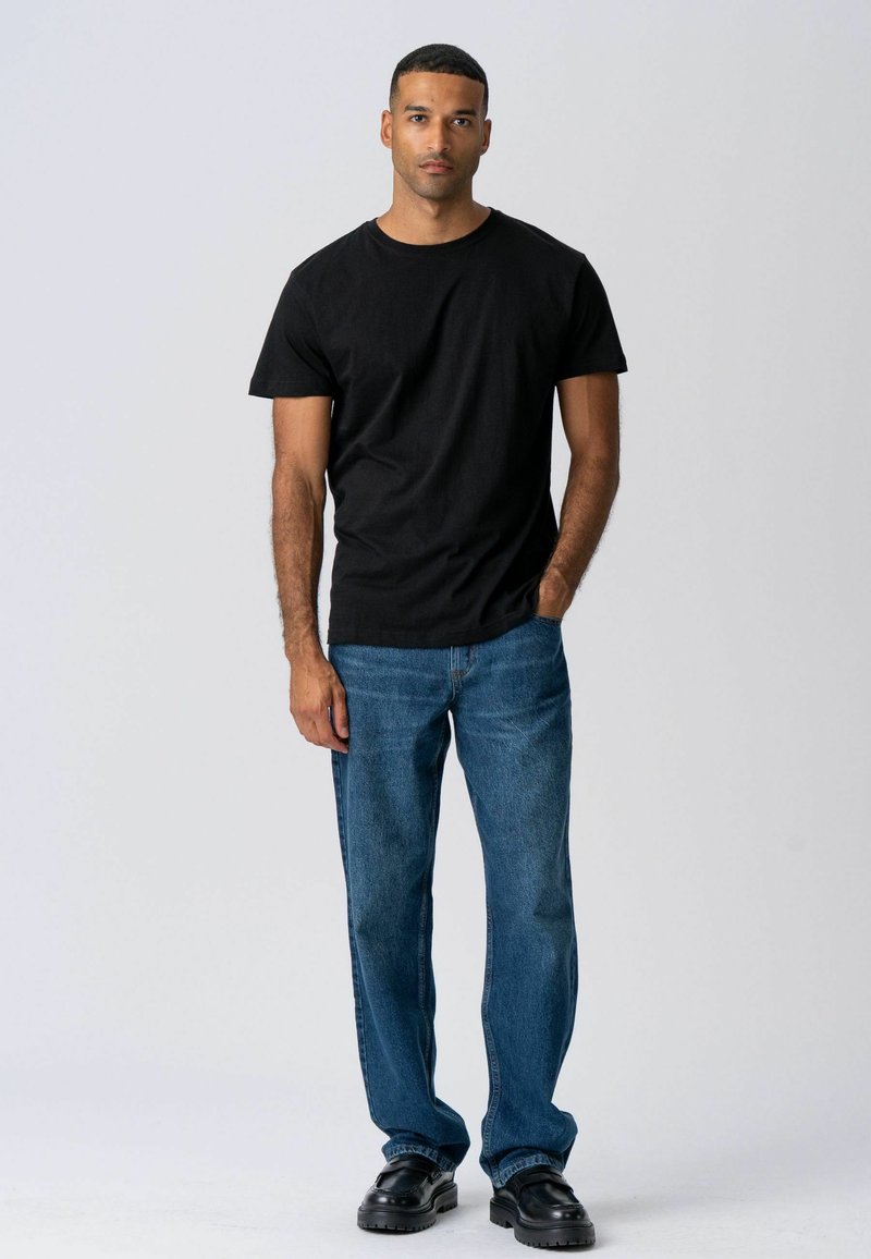 Homme debout contre un fond uni, portant un t-shirt noir, un jean bleu et des chaussures noires, une main dans la poche.