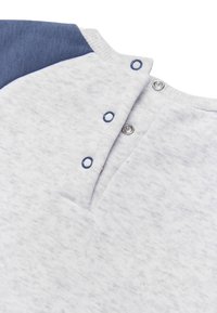 Primer plano de una tela gris claro con un detalle azul en el hombro y tres botones a presión plateados desabrochados a lo largo del escote.