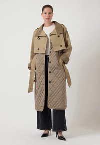 Beiger Trenchcoat, geschichtet über einem gesteppten, gepolsterten Innenstück. Verfügt über Knopfverschluss, weitem Kragen und kombiniert mit schwarzen Hosen mit weitem Bein.