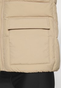 Veste matelassée beige avec fermeture éclair et grande poche à rabat. Présente une texture matelassée et un tissu doux et mat. Détails de coutures apparentes.