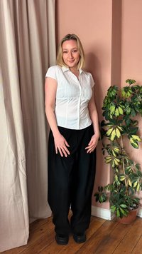 Νεαρή γυναίκα με λευκό πουκάμισο κοντά μανίκια και μαύρο παντελόνι στέκεται χαμογελώντας δίπλα σε μια γλάστρα με πράσινο φυτό μπροστά σε ένα ροδακινί τοίχο.