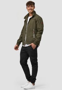 Olivenfarvet bomberjakke med lynlås, ribbede manchetter og to frontlommer. Stylet med en hvid t-shirt, sorte bukser og hvide sneakers.