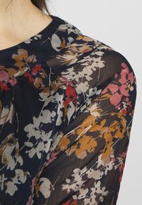 Genomskinlig blommig blus med en marinblå bakgrund och färgglada blomstermönster i röd, beige och guldtoner, med en rundad halsringning.