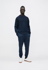 Fiatal férfi zsebre tett kézzel, sötétkék kötött pulóvert, hozzá illő melegítőnadrágot és fehér tornacipőt barna talppal visel.