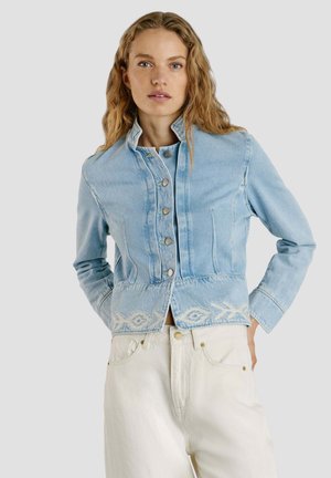 Donna con lunghi capelli mossi che indossa una giacca di jeans azzurro chiaro con bottoni e motivi ricamati bianchi sull'orlo, abbinata a pantaloni bianco sporco.