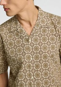 Camicia a maniche corte in tessuto beige caratterizzata da un motivo ripetitivo di forme geometriche marroni e dorate con chiusura a bottoni.